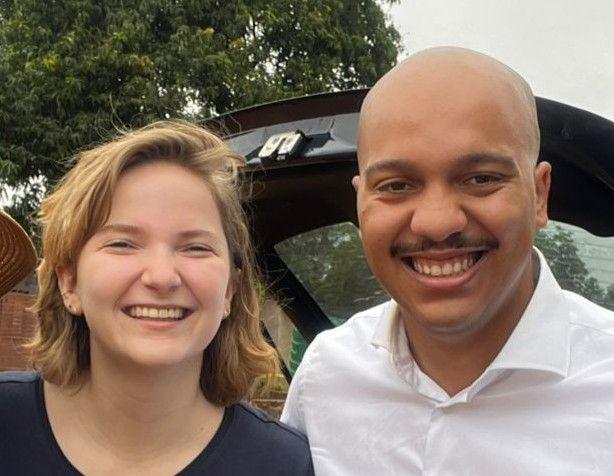 Two people smiling outdoors near an open car trunk with trees in the background.
